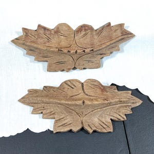 May include: Two light brown wooden decorative carvings shaped like stylized leaves. Each piece features a symmetrical design with carved vein details and a pointed, jagged edge profile. The wood grain is visible across the surface of both items.