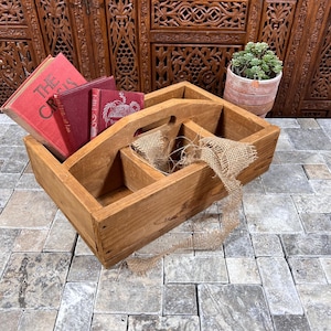 May include: A rectangular wooden caddy with a central handle and three interior compartments. The caddy contains three red hardcover books and a piece of frayed tan burlap ribbon draped over the side of one of the wooden dividers.