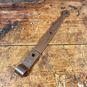 May include: A dark brown, weathered metal strap hinge featuring a long, tapered arm with a rounded, perforated end. The hinge includes a cylindrical barrel section at the base and two additional circular mounting holes along the flat metal arm.