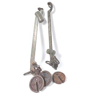 May include: Antique metal balance scale with two arms and several weights. The scale arms are long and slender, with a hook at the top and a mechanism at the bottom. The weights are circular and have a hole in the center. The metal has a weathered appearance.