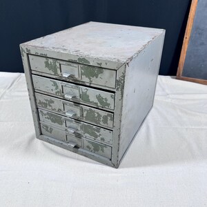 May include: A vintage metal storage cabinet featuring five stacked drawers with distressed olive green paint and worn edges. Each drawer is equipped with a silver-toned metal handle and a label holder, showcasing a weathered industrial aesthetic.