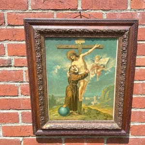 May include: Framed religious artwork depicting a scene of Jesus on the cross, with an angel and a figure embracing him. The artwork is in a dark wood frame, set against a red brick wall.