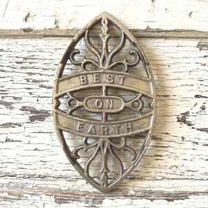 May include: Oval-shaped, antique-style trivet with ornate, openwork design. The trivet is made of a metallic material and features the words "BEST ON EARTH" in raised lettering. The background is a distressed, white-painted wooden surface.
