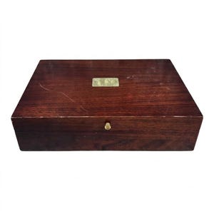 May include: A rectangular dark brown wooden box with a smooth, polished finish. The lid features a centered, blank rectangular brass plaque and a small, circular brass knob centered on the front edge for opening the hinged lid.