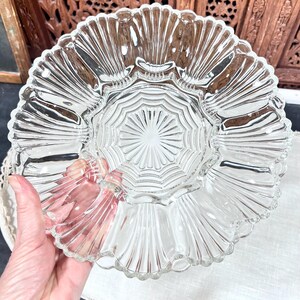 May include: Clear glass serving dish with a scalloped edge and a central, starburst-patterned design. The dish has a fluted, fan-like pattern radiating from the center. The dish is held by a person's hand.