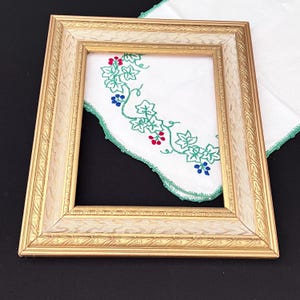May include: A rectangular, gold-toned picture frame with a decorative border. The frame surrounds a white cloth with green, red, and blue embroidered floral details. The cloth has a green scalloped edge. The frame is empty.