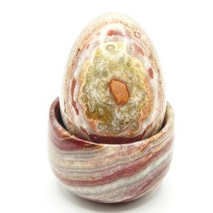 May include: A decorative egg-shaped stone sculpture in a matching bowl. The egg is primarily beige, green, and brown with a speckled pattern. The bowl has horizontal bands of red, white, and gray.