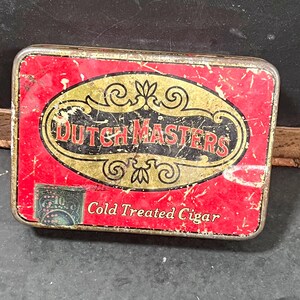 May include: A rectangular, vintage Dutch Masters cigar tin. The tin has a red background with a gold and black oval design in the center, featuring the words "Dutch Masters." The text "Cold Treated Cigar" is also visible. The tin shows signs of wear.