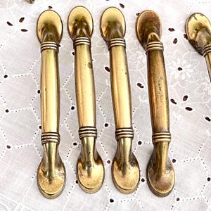 May include: A collection of vintage brass drawer pulls. Each pull features a long, cylindrical handle with decorative rings and a rounded end. The pulls have a warm, golden hue and show signs of age.