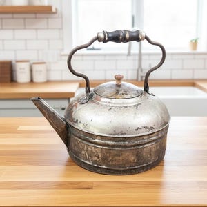 May include: A weathered metal tea kettle with a distressed silver finish, featuring a dark wooden handle with silver accents, a matching lid with a small wooden knob, and a wide spout. The kettle has a cylindrical body with horizontal ridges.