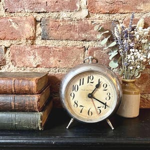 May include: A silver-colored, round alarm clock with a cream-colored face and black numerals. The clock is labeled "The Autonomic." Beside the clock are three antique books and a small vase with dried flowers. The background is a brick wall.
