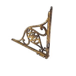 May include: A triangular metal shelf bracket featuring a distressed gold finish over a dark base. The bracket has an ornate, openwork scroll design with a central lattice pattern and mounting holes at the top and bottom ends of the vertical spine.