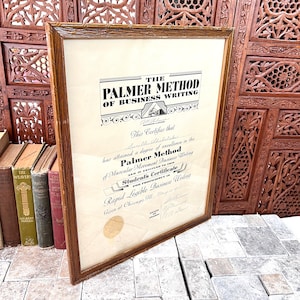 May include: A framed certificate titled The Palmer Method of Business Writing, featuring black text on an off-white background, a gold seal, and handwritten details. The document is enclosed in a brown wooden frame with a textured, carved border.