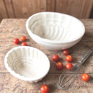 May include: Two white ceramic bundt cake molds with a swirl design sit on a wooden surface, alongside a whisk and several red cherry tomatoes. The molds are different sizes and have a vintage aesthetic.