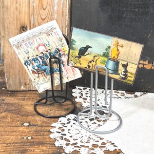 May include: Two metal paperclip-shaped card holders, one black and one silver, each featuring a circular base. The black holder displays a vintage postcard of a formal gathering, while the silver holder displays a postcard with a crow, dog, and duck.