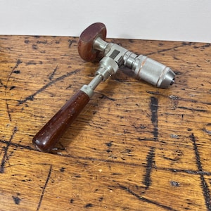 May include: A vintage hand drill featuring a polished metal body with a knurled chuck and a dark brown wooden handle. The tool includes a rounded wooden palm rest at the top, connected to a central metal shaft and a perpendicular handle grip.