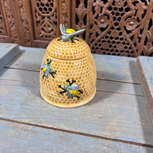 May include: A ceramic honey pot with a textured honeycomb pattern in shades of yellow and tan. The pot features a removable lid topped with a sculpted bee, and two additional bees are painted on the sides with grey wings and yellow bodies.