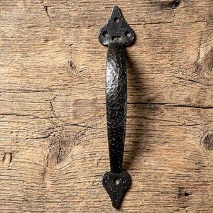 May include: A black, textured, wrought iron door pull with a hammered finish. The handle has a teardrop shape at each end with a central grip. The door pull is set against a rustic wooden background.