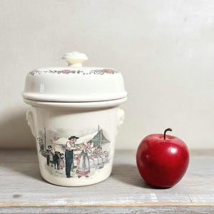 May include: A cream-colored ceramic jar with a lid, featuring a floral design and a scene of people at a market. A red apple sits beside the jar. The jar has two small handles.