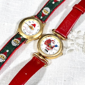 May include: Two gold-tone wristwatches with round faces. One watch has a green strap with Santa Claus faces and a white dial reading Merry Christmas. The other watch has a solid red strap and a white dial featuring a full-body Santa Claus graphic.