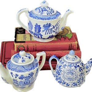May include: Three ceramic teapots with blue and white floral and landscape designs. The teapots are stacked on top of each other, with the top teapot featuring a detailed landscape scene. The teapots are placed on top of a stack of red books.