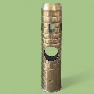 May include: A tall, brass-colored metal umbrella stand with a cylindrical shape. The stand features a series of rectangular cutouts with mesh, a large circular opening, and a small door. The design resembles a tower.