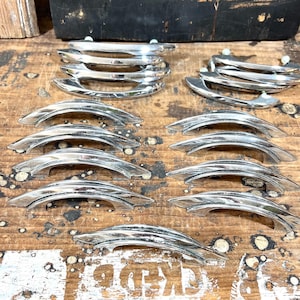 May include: A collection of twelve polished chrome cabinet pulls arranged in rows. Each handle features a curved, arching design with a smooth, reflective metallic finish and two mounting points on the underside for attachment to furniture.