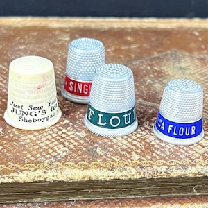 May include: Four vintage thimbles with text. One thimble is cream-colored with the text "Just Sew You JUNG'S for Sheboygan". The other three thimbles are silver-colored with colored bands that read "SING", "FLOU", and "CA FLOUR".