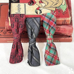 May include: Three vintage bow ties in a row. One is burgundy with a repeating pattern, the second is gray with stripes, and the third is a red, green, and white plaid. The bow ties are resting on top of a stack of old books.