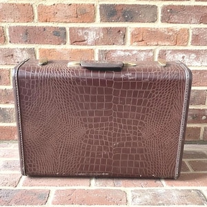 May include: A rectangular brown suitcase featuring a textured crocodile skin pattern across its exterior surface. The luggage includes a dark brown top handle, metallic gold-toned hardware latches, and reinforced edges along the entire perimeter.
