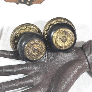 May include: Two round door knobs featuring smooth black centers and ornate, circular brass-toned metal plates. Each knob has a decorative, sunburst-patterned brass inlay on the front face, showcasing intricate detailing and a metallic finish.