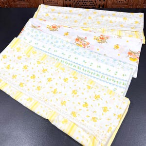 May include: A stack of folded fabric pieces featuring various floral patterns. The textiles include white backgrounds with yellow, orange, and light blue flower prints, accented by yellow striped borders and delicate white lace trim details.