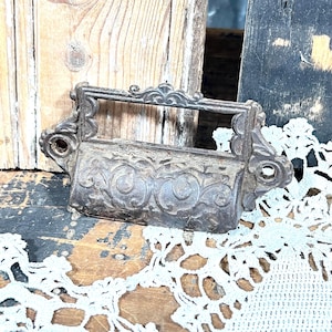 May include: A dark, weathered metal drawer pull featuring an ornate, raised scrollwork pattern across its front surface. The hardware includes a rectangular label holder frame at the top and two circular screw holes on the side mounting flanges.