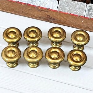 May include: Eight brass cabinet knobs with a classic design. Each knob features a rounded top with a recessed center and a decorative base. The knobs are arranged in two rows of four on a white wooden surface.