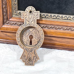 May include: Ornate bronze-colored keyhole cover with intricate floral and geometric designs. The cover features a cross-shaped top and a circular center with a keyhole. The design includes detailed carvings of leaves, flowers, and symmetrical patterns, suitable for antique decor.
