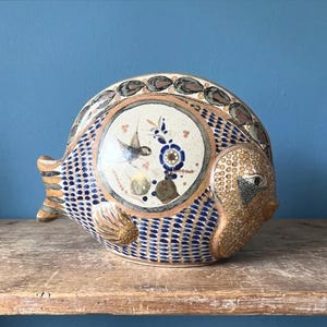 May include: A ceramic fish sculpture with a round body and a detailed design. The fish features a central circular design with a bird and floral elements. The body is primarily blue with a dotted pattern, and the head and tail have textured details.