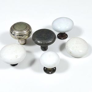 May include: An assortment of vintage door knobs in various styles. The collection includes knobs in silver, black, and white, with a mix of materials like metal and ceramic. The knobs are arranged on a white surface.