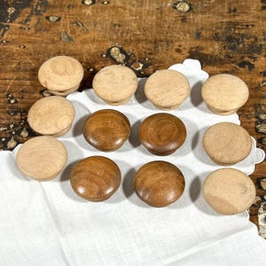 May include: A collection of twelve round wooden knobs arranged in a grid. The set includes eight light-colored natural wood knobs and four darker, stained brown wood knobs. Each knob features a smooth, domed top and a circular base for mounting.
