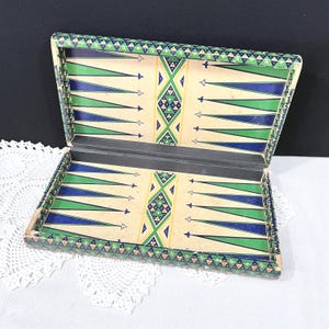 May include: Open backgammon board with a cream-colored interior featuring blue and green triangles. The board's edges are decorated with a repeating geometric pattern in green and blue. The board is open, revealing the playing surface.