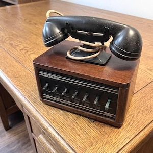 May include: Vintage black telephone with a handset and a wooden base. The base features several switches and the text "BE VISOR FOR OFFICE". The phone is sitting on a wooden desk.