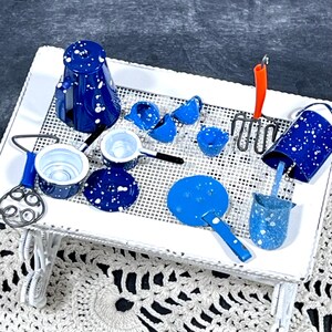 May include: A miniature kitchen set featuring a dark blue coffee pot, a small saucepan, a lid, a frying pan, a bucket, a rake with an orange handle, and a whisk, all decorated with white paint splatters on a white metal mesh tray table.