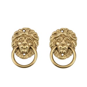 May include: Two antique brass lion head door knockers. Each knocker features a detailed lion's face with a ring-shaped handle. The knockers are a warm gold color and are set against a white background.