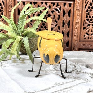 May include: A yellow ceramic bee-shaped container with black stripes and large black eyes. The container has a leaf-shaped lid and sits on a metal stand. A small wooden spoon rests inside. A green plant is in the background.