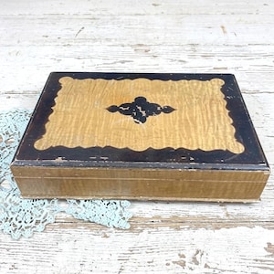 May include: A rectangular wooden box featuring a light tan, wavy-grained wood top bordered by a dark brown, scalloped frame. A dark brown, four-pointed decorative emblem is centered on the lid. The sides of the box are light tan wood.