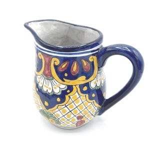 May include: A ceramic pitcher with a dark blue base and a curved handle. The pitcher is decorated with yellow, red, and white floral patterns. The interior is white. The pitcher has a spout for easy pouring.