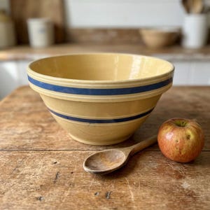 May include: A large, round, yellow ceramic mixing bowl featuring two horizontal dark blue stripes encircling the exterior. The bowl has a wide, flared rim and a smooth, glossy finish, presenting a classic design for kitchen use and food preparation.