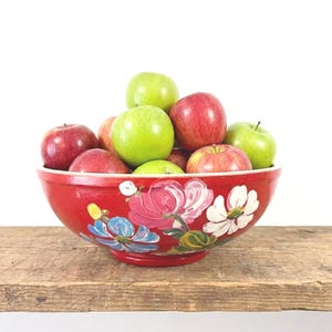 May include: A vibrant red bowl filled with a variety of apples. The bowl is adorned with hand-painted floral designs in pink, white, and blue. The apples are a mix of red and green.