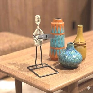 May include: A decorative table arrangement featuring a metal and woven figure holding a candle holder, alongside three ceramic vases. The vases are in shades of orange, teal, blue, and yellow, with various patterns. The arrangement sits on a wooden table.