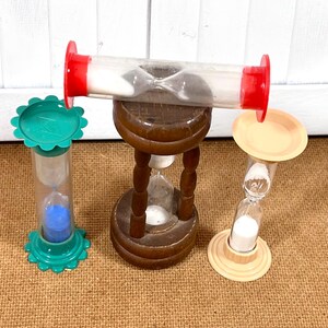 May include: Three hourglasses of varying designs and colors. One is a clear tube with red end caps, another is a wooden hourglass, and the third is green and clear with blue sand. The hourglasses are on a brown surface.