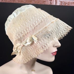 May include: A vintage cream-colored lace bonnet with a delicate design. The bonnet features a brim with a scalloped edge and is adorned with a satin ribbon and a small pink bow. The hat is displayed on a mannequin head.